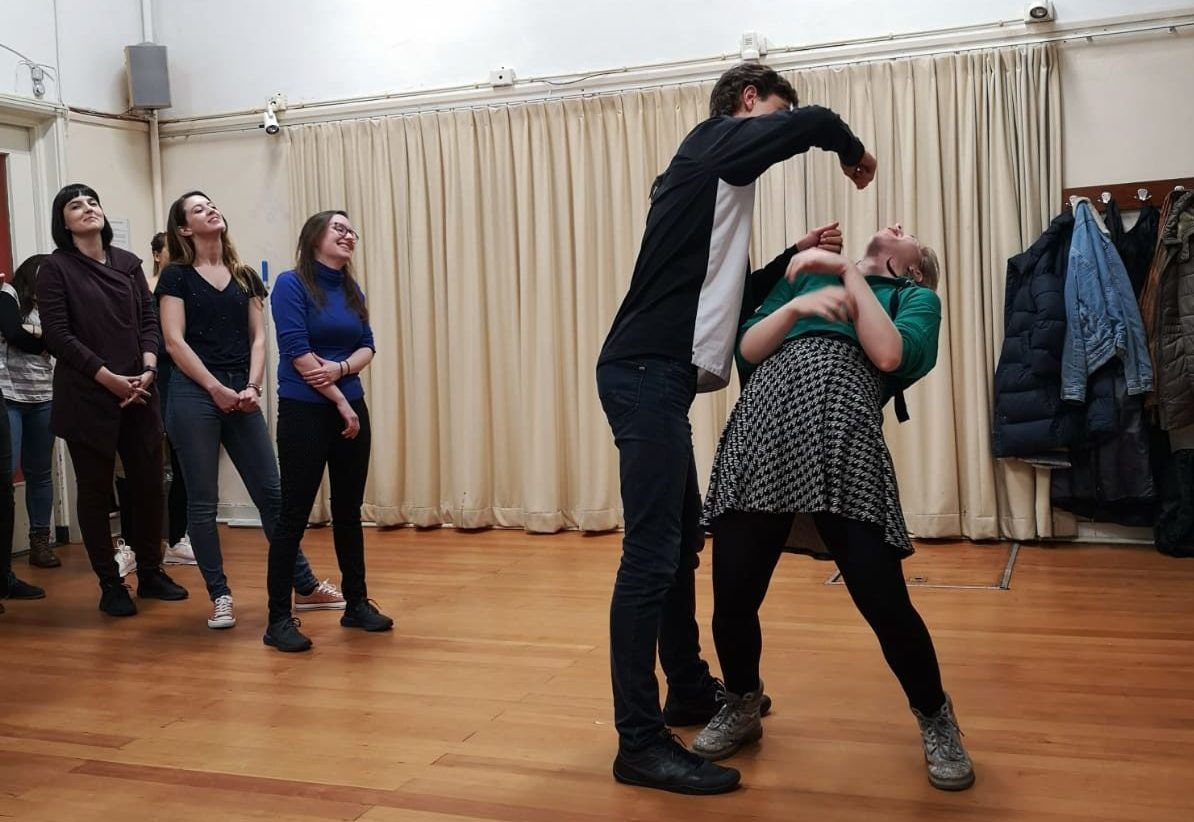 Improv Class Act Attack. A person has their arm hanging on top of another person's face, like trying to feed or punch them. The second person, clearly a girl, leans backwards
