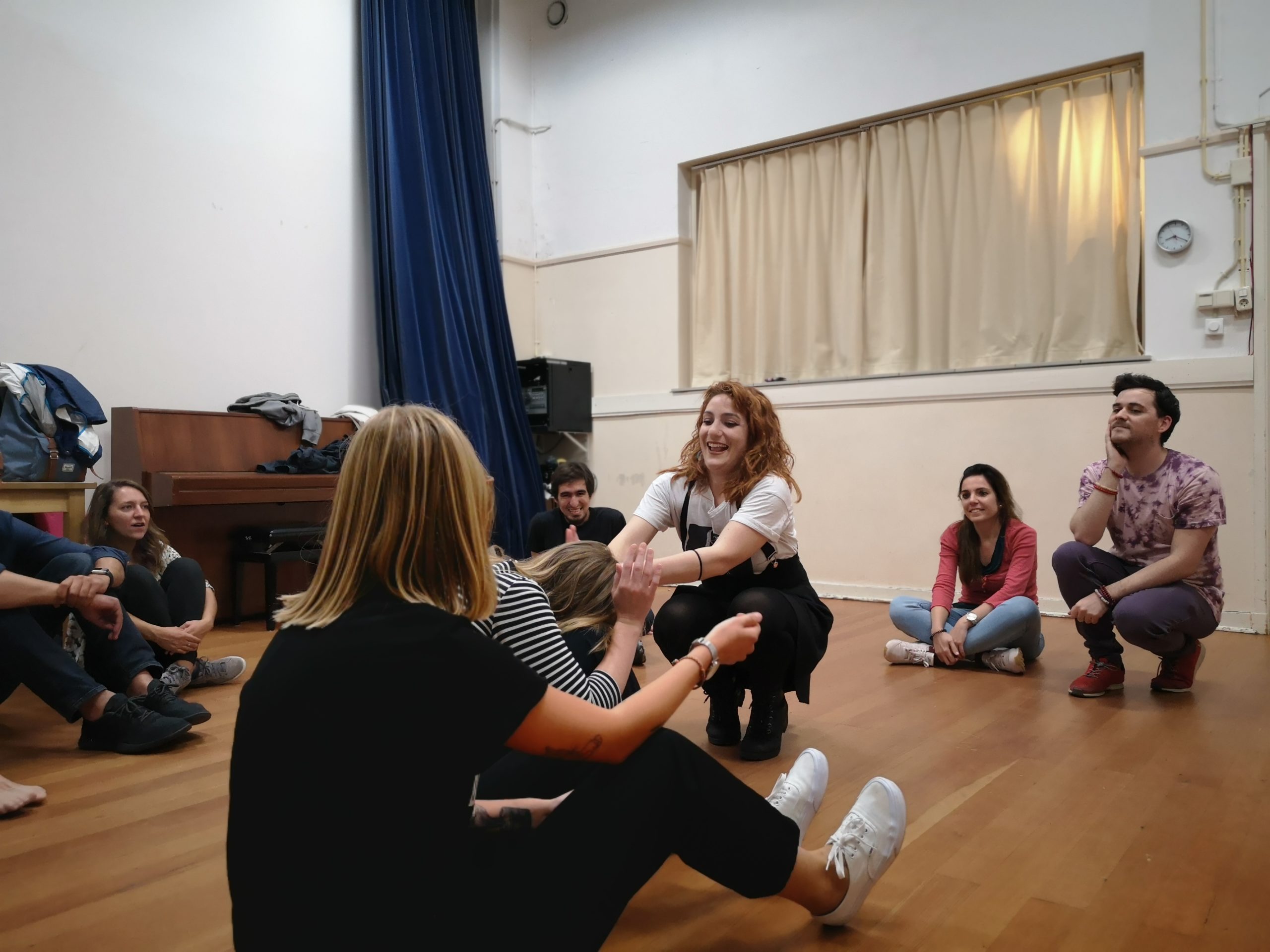 From the improv and physical class. A group sits in a circle. Three women are in the middle, two sitting, one kneeling and laughing, reaching her hands to one of the others.
