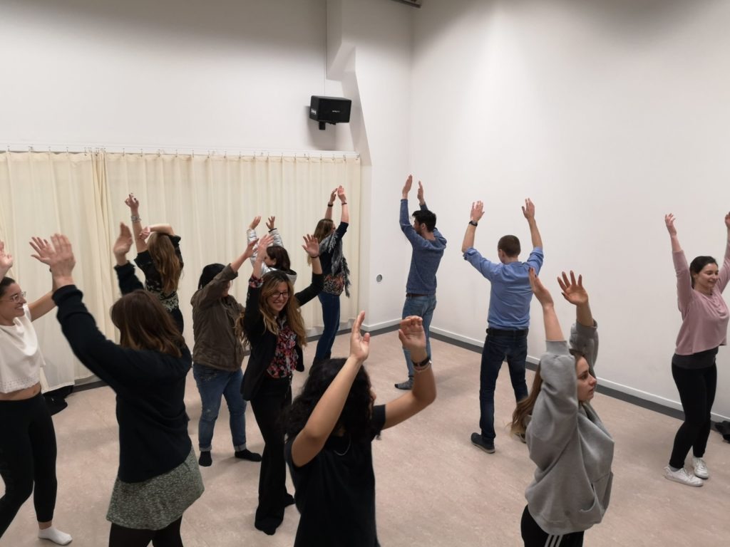 Photo from a class. A group of people are scattered around the room, with their hands in the air