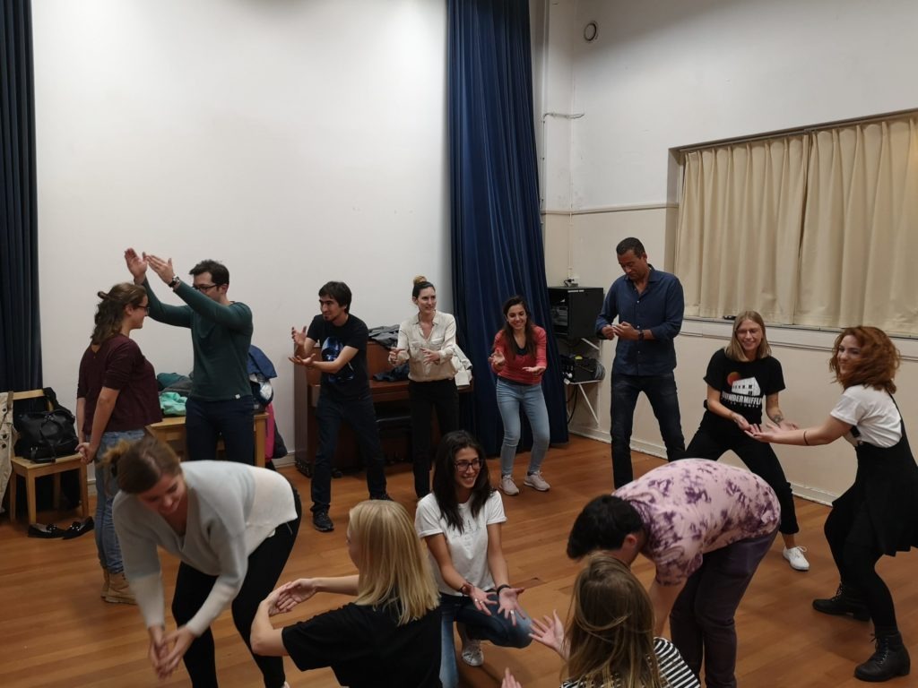 From the improv and physical class. A group of people do an exercise. Some are sitting, some kneeling, some standing, everybody is moving their arms differently.