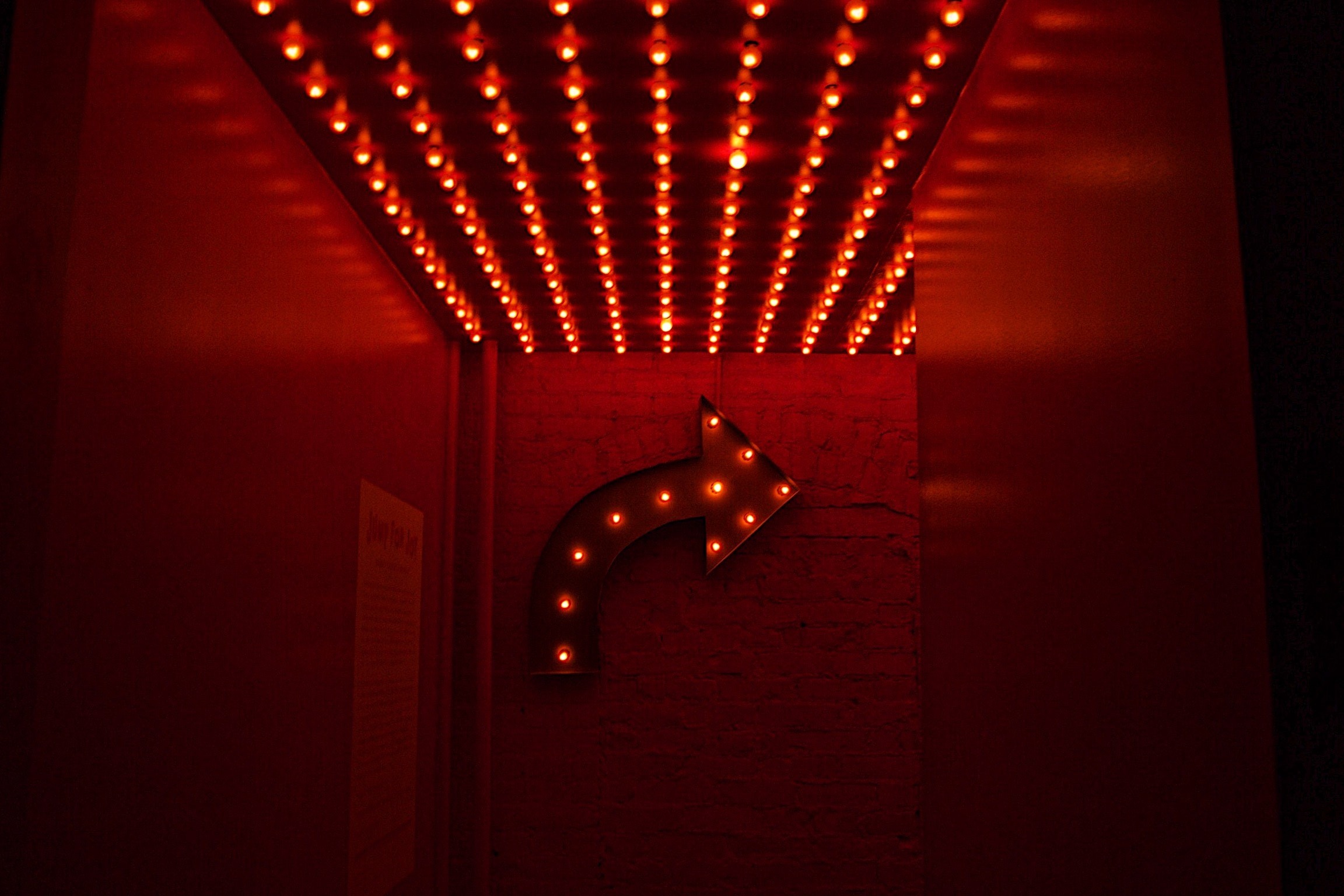 red photo. A series of light bulbs for, the shape of an arrow pointing right. A series of red lightbulbs are placed on the ceiling as well.
