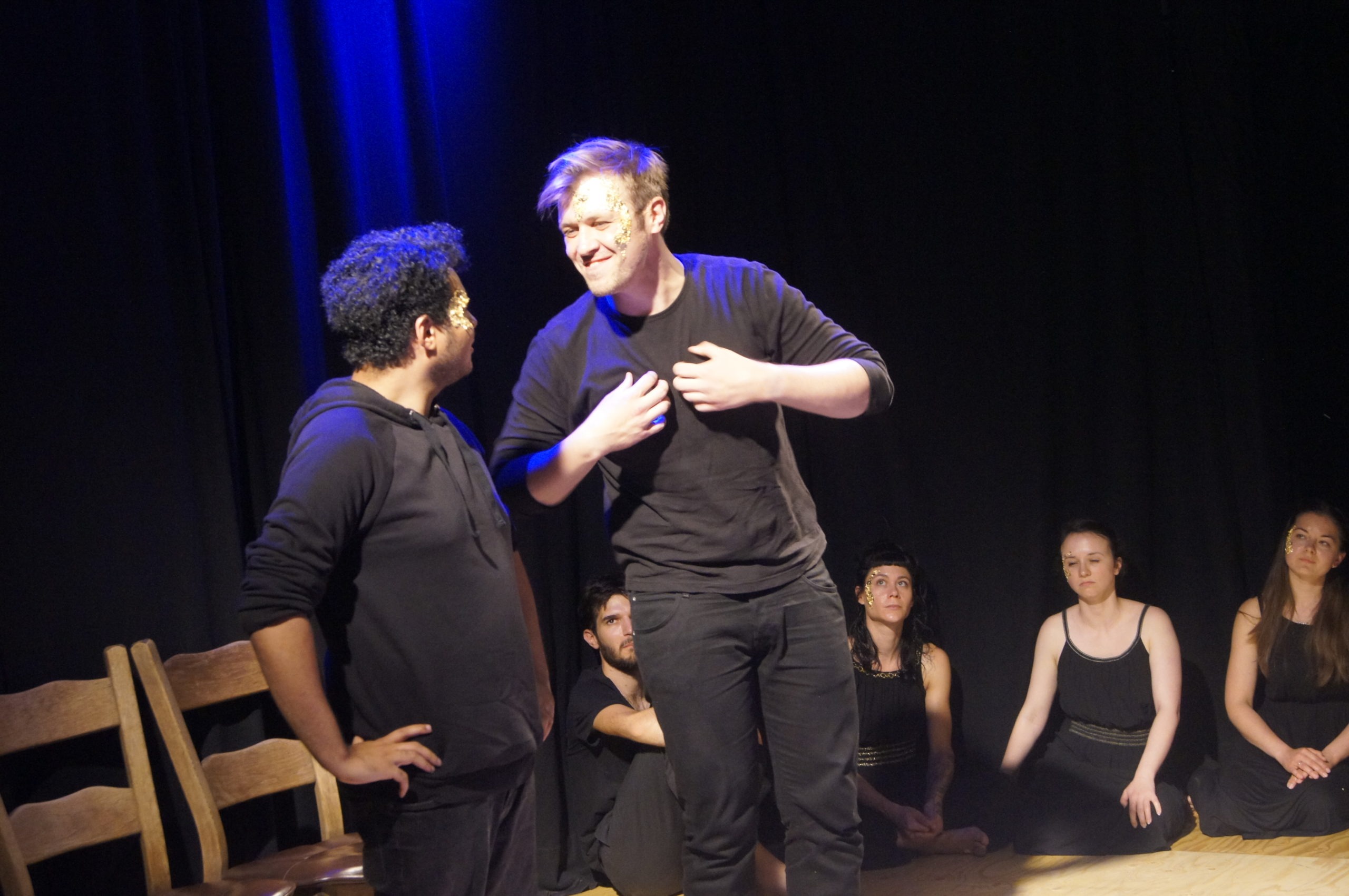 Photo from the theatre performance Keeping up with the Greeks. Two male actors on stage, standing, wearing black clothes, talking, one of them grins and holds his chest with his hands. Behind them, four actors on the floor, black clothes, golden flakes on their face