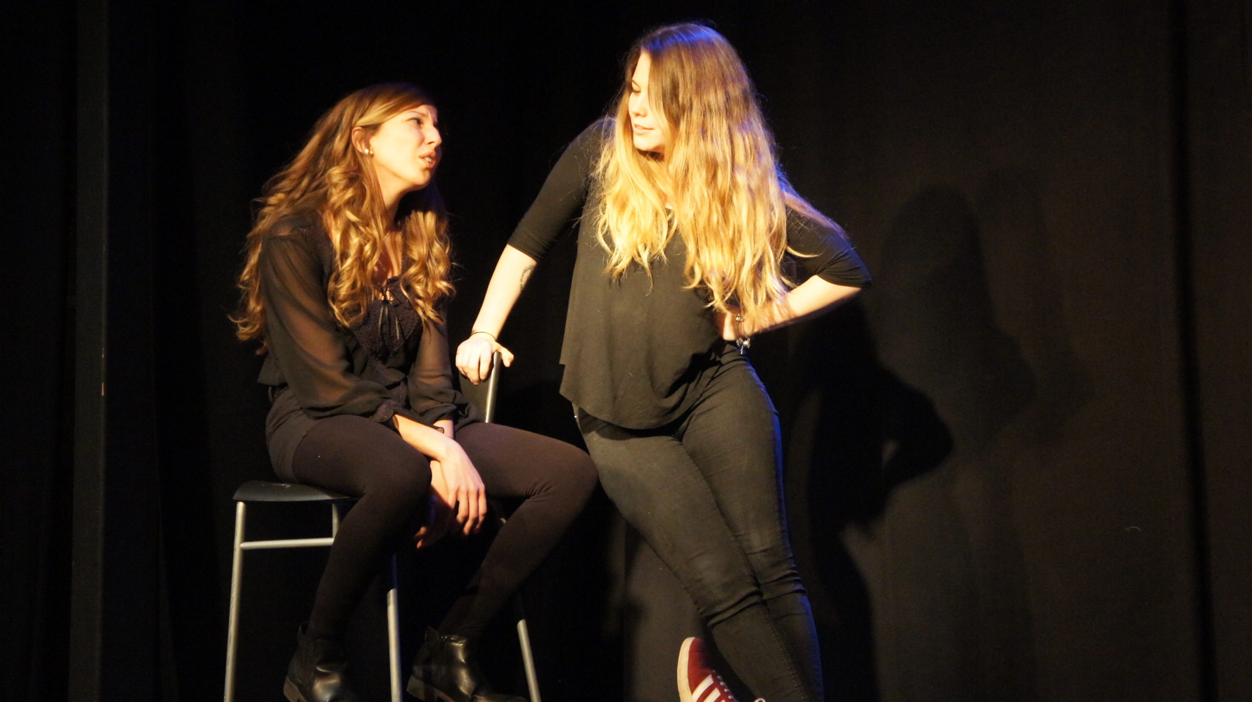 Act Attack's improv and physical show. Two girls on stage, wearing black. One sitting on a bar stool, the other leaning on her. They're talking