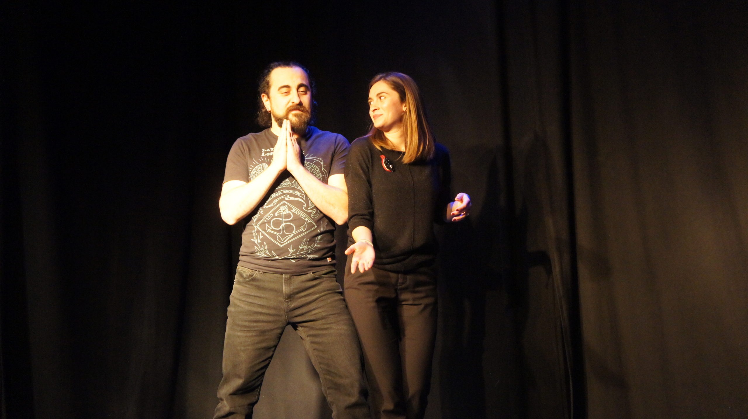 Act Attack's improv and physical show. A man and a woman on stage wearing black. The woman looks at the man showing him something on the floor, he has his hands placed in front of his heart, like praying