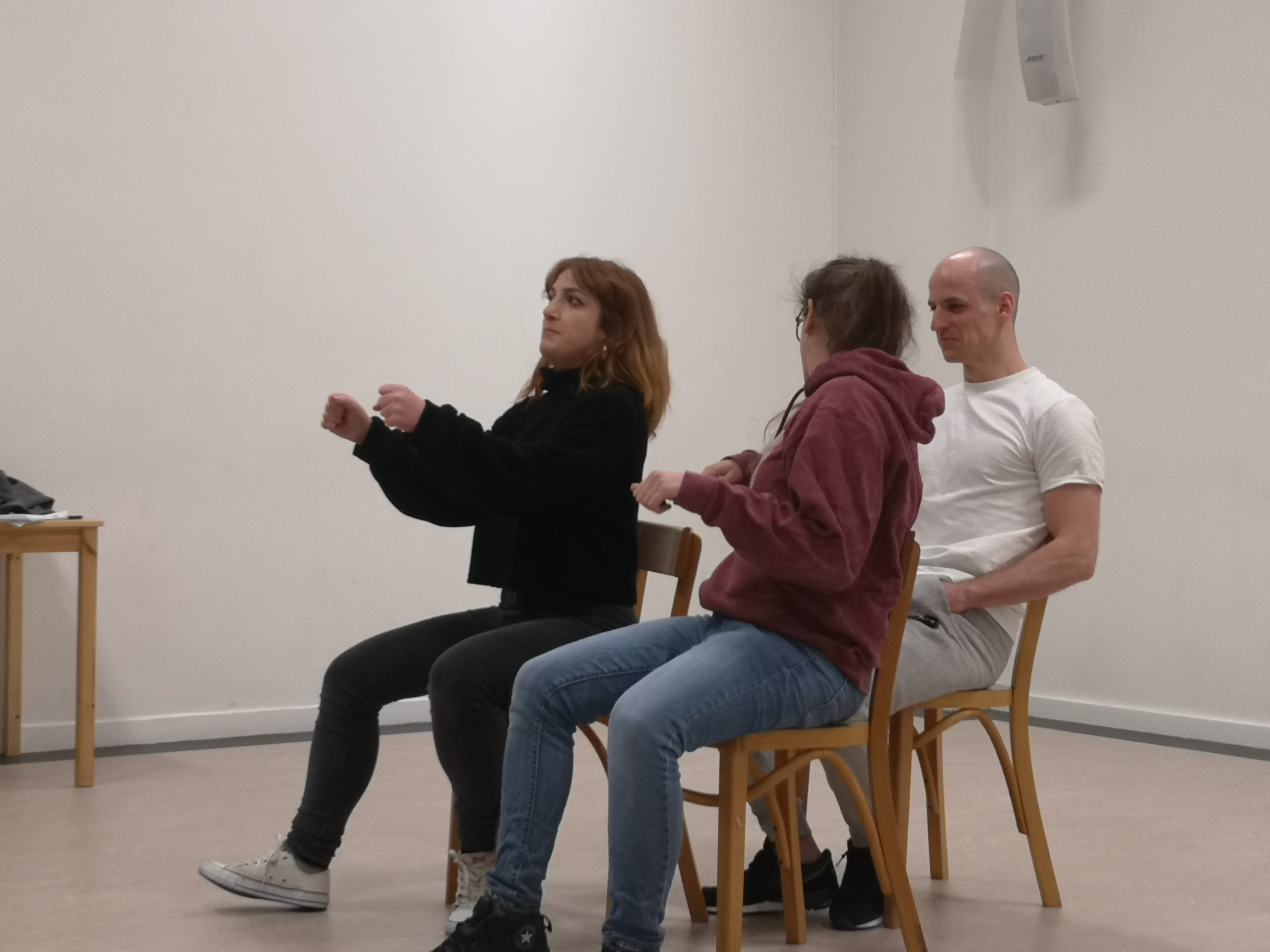 Acting class Act Attack. Three people are sitting on chairs, two at the front one at the back. The front right person, a girl, pretends to drive