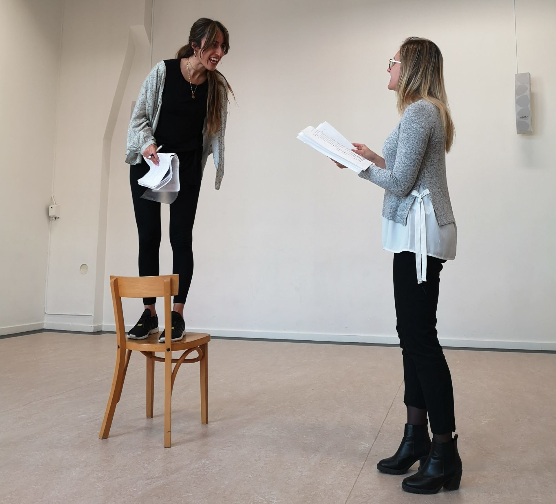 Acting Class Act Attack. Two women read lines, one is standing on the chair, laughing or screaming, the other one is standing up.