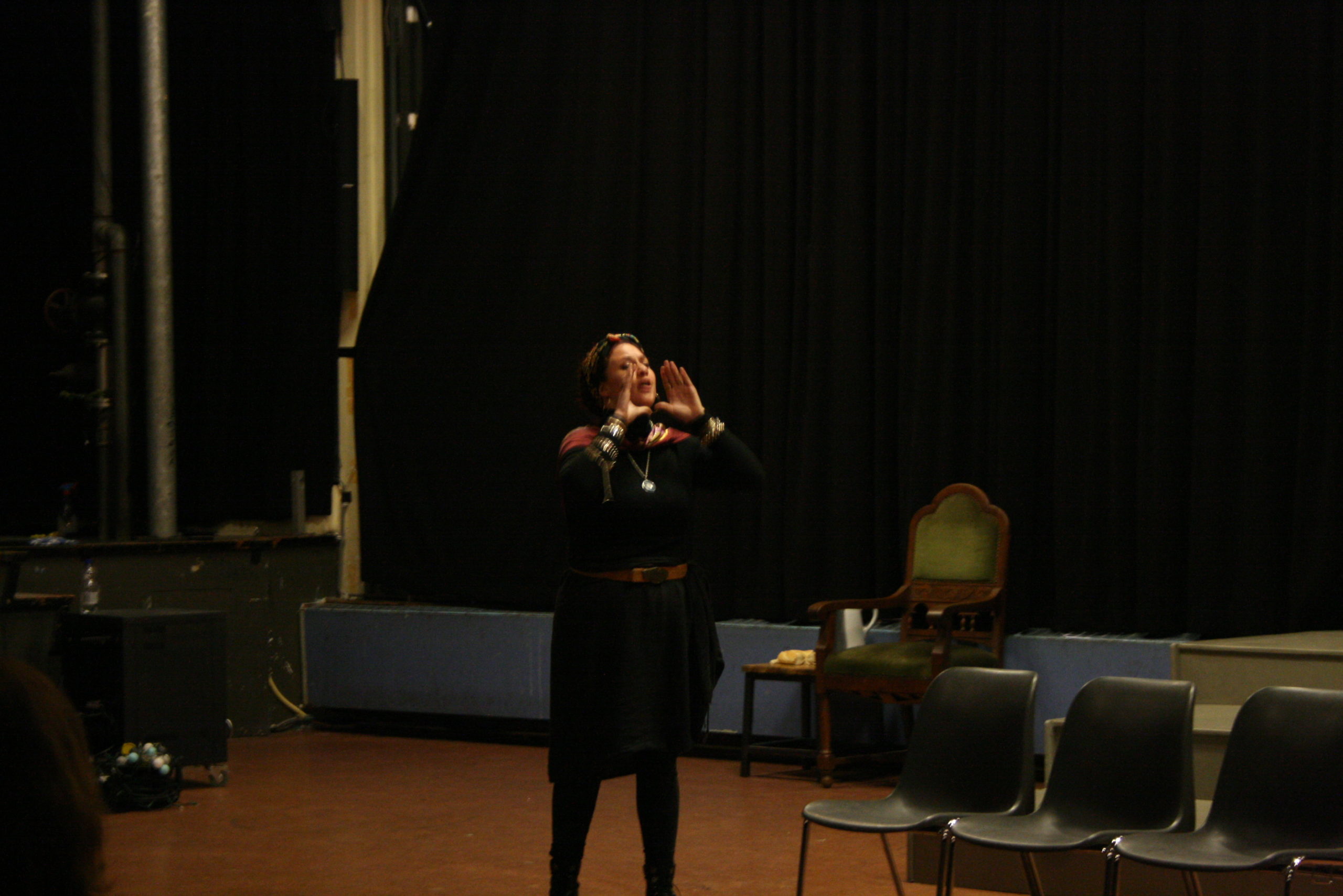 Photo from the show "Skin of our Teeth". A female actor on stage, dressed mostly in black, has her hands in front of her mouth like she's calling someone