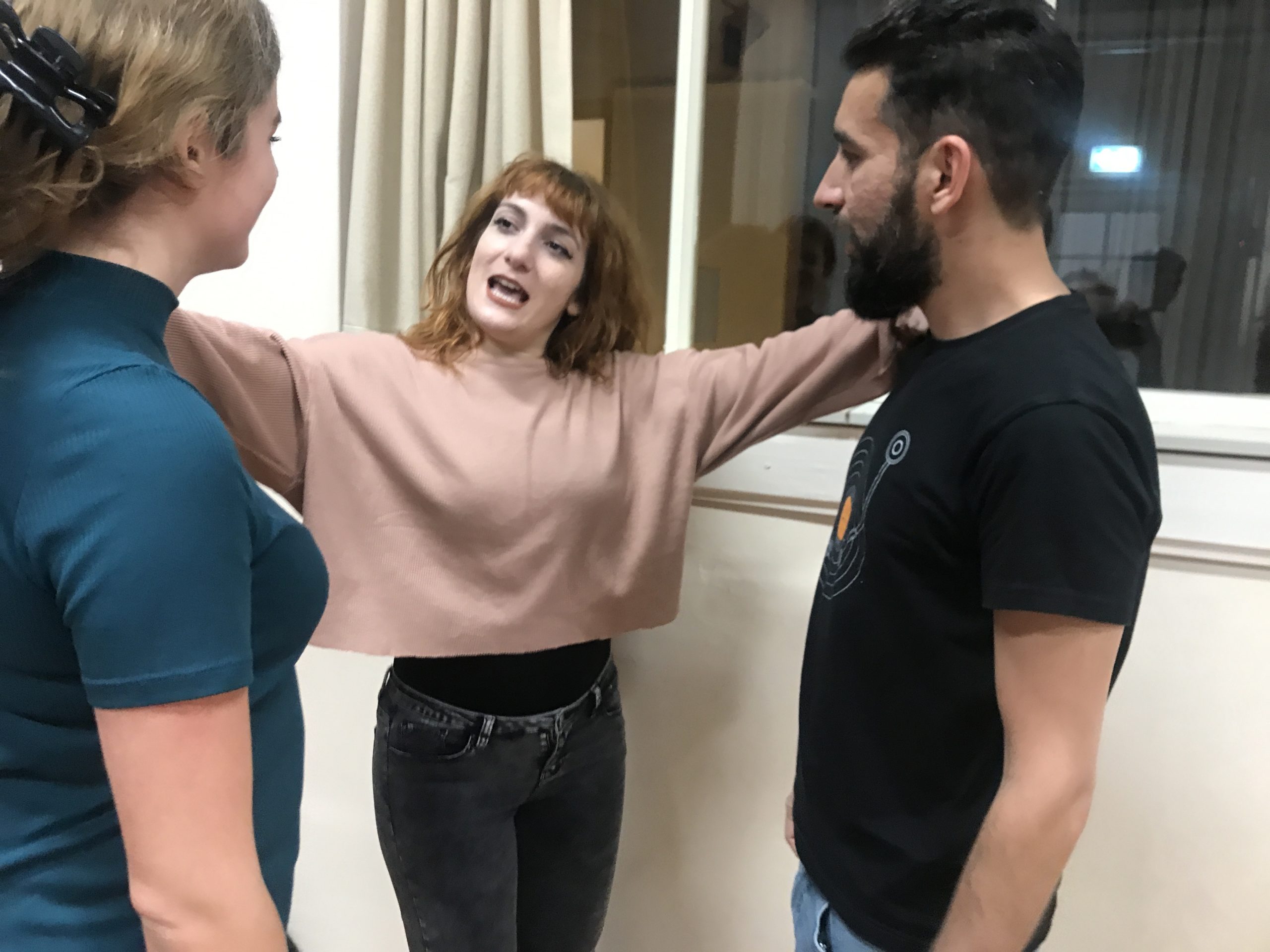 Act Attack's Improv Class. A woman in the middle reaches her arms to touch a woman to her right and a man to her left. She looks at the woman on her right, speaking to her