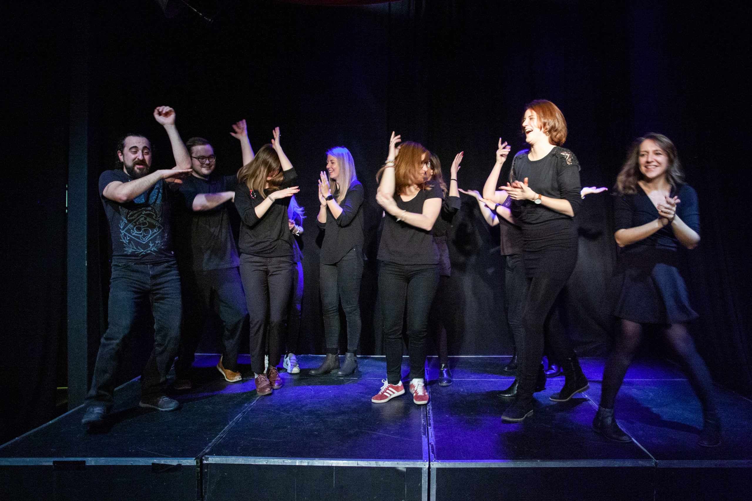 Act Attack's improv and physical show. A group of people on stage wearing black move their arms. Some have their arms placed in the shape of an L