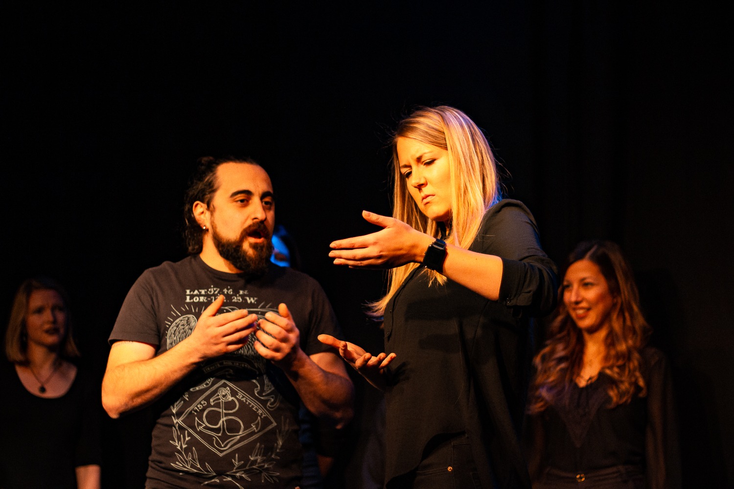 Act Attack's improv and physical show. A group of people on stage wearing black clothes. A man with beard speaks to a blonde woman who pretends to hold something in her arms.