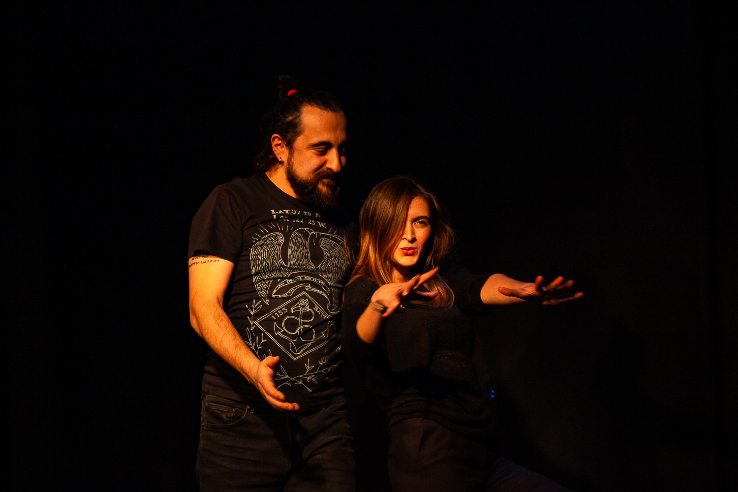 Act Attack's improv and physical show. A man and a woman on stage wearing black. The woman is posing or reaching her arms to show something, the man looks at her grinning