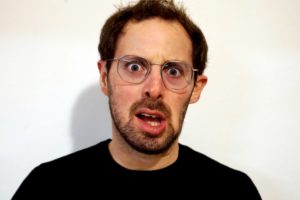 Act Attack teacher Isaac Lester Simon. Man, white, brown hair, blue eyes, beard, black top, grey metal glasses. He looks shocked but in a funny way, his lower lip drops to the side